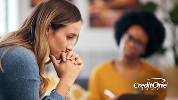 A young woman rests her chin on her hands as if she’s contemplating a crisis while another woman, who is perhaps her therapist or credit counselor, looks on with a clipboard.
